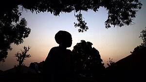 Eine Person steht in der Natur bei aufgehender Sonne. Die Umgebung und die Person sind schattiert, nur der Himmel leuchtet farbig und erzeugt eine melancholische Stimmung.