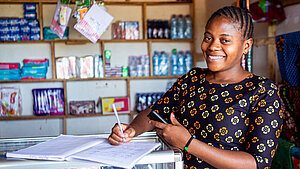 Vertrauensgarantie Eine Frau steht an einem Tresen und hält einen Stift in der Hand. Vor ihr liegt ein Heft. Im Hintergrund sind Regale mit Lebensmitteln und Stoffen zu sehen. Sie lächelt in die Kamera.
