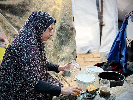 Mutter aus Gaza bereitet Essen zu Eine Mutter aus Gaza bereitet im Freien Essen zu. Um sie herum sind Zelte und Schutt.