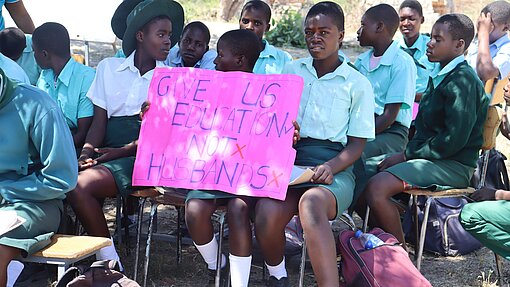 Eine Gruppe von Schüler:innen sitzt auf Stühlen und hört zu. Eine Schülerin hält ein leuchtend pinkes Plakat, auf dem steht: "GIVE US EDUCATION, NOT HUSBANDS". Das Plakat ist in großen, bunten Buchstaben geschrieben. Die Schüler:innen tragen Schuluniformen.