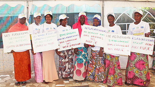 Junge Frauen und Mädchen in traditioneller Kleidung und weißen Plan T-Shirts stehen nebeneinander unter einem Zelt. Sie halten Schilder mit französischen Botschaften gegen weibliche Genitalverstümmelung.