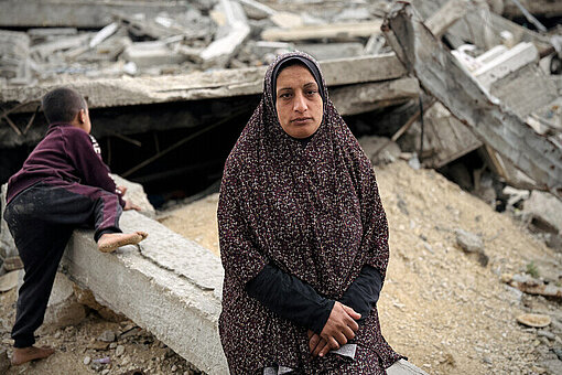 Suhad sitzt inmitten der Überreste ihres zerstörten Zuhauses Eine Mutter aus Gaza steht vor den Trümmern ihres Hauses. Hinter ihr klettert ein kleiner Junge gerade auf ein Trümmerteil.