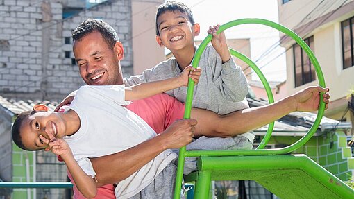 Zwei Jungen spielen mit ihrem Vater ausgelassen auf einem Spielplatz Zwei Jungen spielen mit ihrem Vater ausgelassen auf einem Spielplatz. Gerade klettern sie auf einer grünen Rutsche herum.