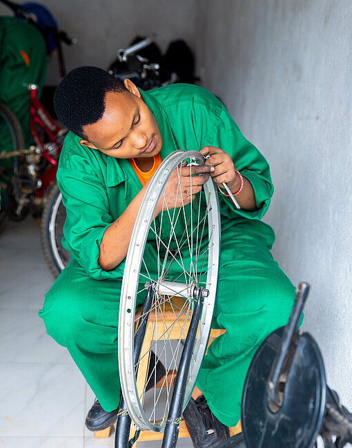 Eine junge Frau im Arbeitsanzug sitzt auf einem Hocker und repariert ein Vorderrad.