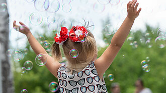 Ein Mädchen steht mit dem Rücken zur Kamera zwischen Seifenblasen und hält die Hände in den Himmel