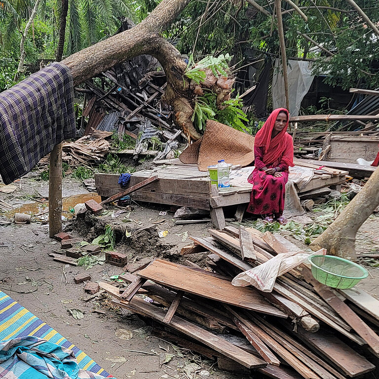 Frau sitzt neben zerstörten Hütten und Häusern umgeben von Wald