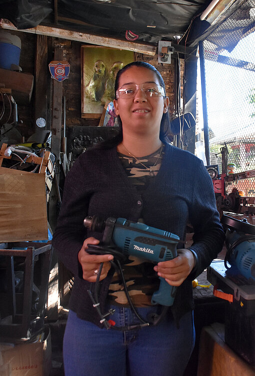 Frau mit Schutzbrille in ihrer Werkstatt mit Bohrmaschine in der Hand blickt in die Kamera