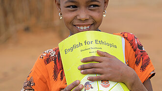 Schulmaterialien für bedürftige Kinder Ein Mädchen hält ein Englisch-Heft in der Hand und lächelt.