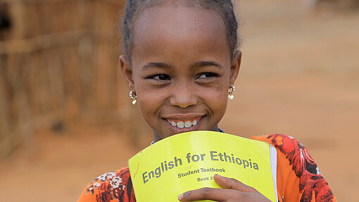 Schulmaterialien für bedürftige Kinder Ein Mädchen hält ein Englisch-Heft in der Hand und lächelt.