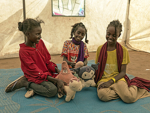 Drei jugendliche Mädchen sitzen in einem großen Zelt auf dem Boden und halten Plüschtiere in der Hand
