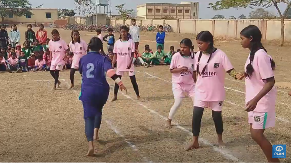 Eine Gruppe von Mädchen in rosa Trikots steht zusammen auf einem Spielfeld, ein Mädchen in blauem Trikot läuft auf sie zu