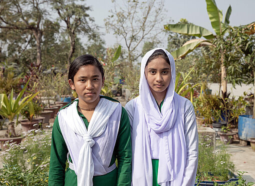 Zwei Mädchen in grünen Schuluniformen stehen zwischen Palmen und Beeten