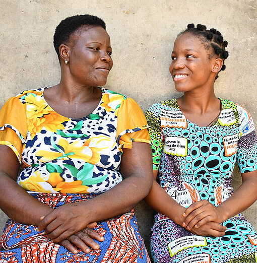 Eine Mutter und Tochter aus Benin in bunter Kleidung sitzen auf einer Bank vor ihrem Haus