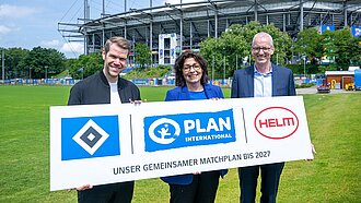 Drei Personen stehen vor dem Stadion und halten ein Banner in der Hand mit den Logos vom HSV, Plan International und der HELM AG