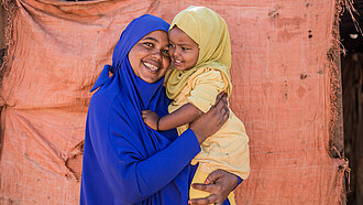 Cawo hält ihre einjährige Tochter im Arm, Cawo lächelt und ihre Tochter schaut sie lächelnd an.