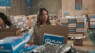 Eine Mitarbeiterin von Plan-International steht in einem Lager und packt Kartons, auf denen ein Plan-Logo drauf ist