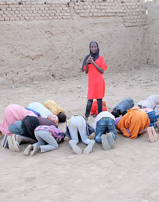 Ein Mädchen steht in der Mitte eines Kreises aus Kindern, die auf dem Boden knien und den Kopf auf den Boden gelegt haben, um ihre Augen zu verdecken