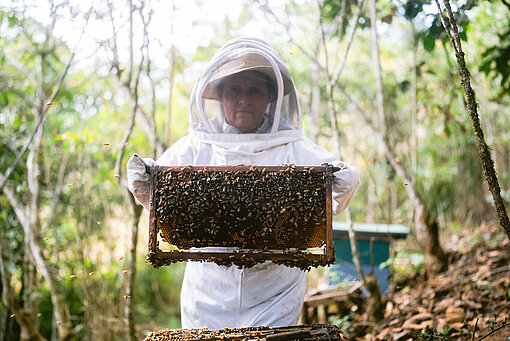 Eine Frau in einem Imkeranzug hält eine Bienenwabe in den Händen und schaut in die Kamera