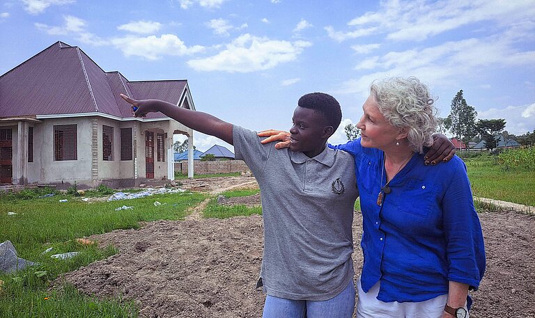 Eine junge tansanische Frau zeigt auf ein Haus, neben ihr steht eine ältere weiße Frau und schaut ihrem Blick hinterher