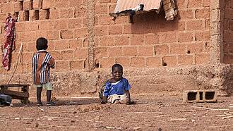 Ein Junge sitzt vor einem Lehmhaus, ein weiterer Junge steht neben ihm