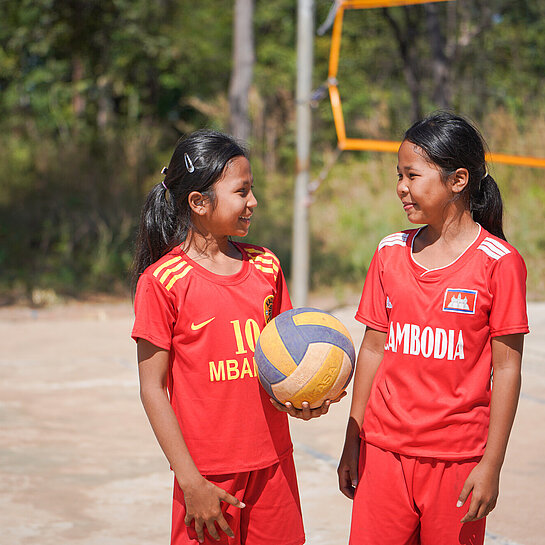 Zwei Mädchen in roten Trikots stehen auf einem Volleyballplatz, sie schauen sich an, eine von beiden hält einen Volleyball in der Hand