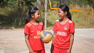 Zwei Mädchen in roten Trikots stehen auf einem Volleyballplatz, sie schauen sich an, eine von beiden hält einen Volleyball in der Hand