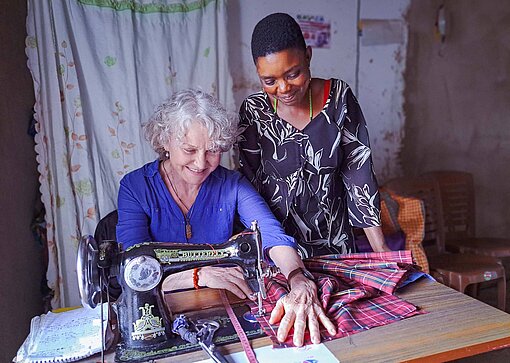 Eine ältere weiße Frau sitzt an einer Nähmaschine und näht ein Stück Stoff, eine junge tansanische Frau steht daneben und schaut zu