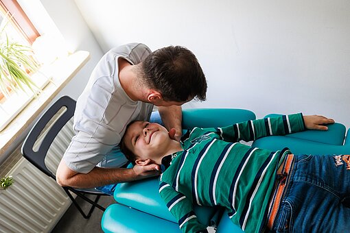 Ein anderer Physiotherapeut behandelt einen Jungen, der auf einer Liege liegt, am Nacken. Der Junge lächelt.