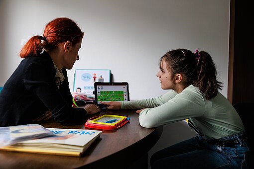 Eine Frau sitzt mit einem Mädchen an einem Tisch, sie schauen auf ein iPad, das Mädchen drückt auf ein Symbol auf dem Gerät.