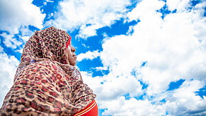 Eine junge Frau ist seitlich von unten fotografiert, hinter ihr sieht man weiße Wolken auf blauem Himmel. Sie schaut seitlich in den Himmel.