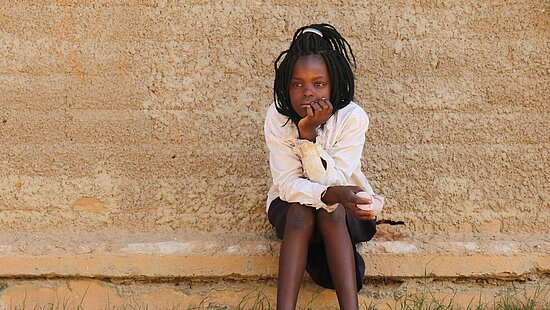 Ein Mädchen sitzt auf einem Vorsprung vor einer Lehmwand und hat den Kopf auf die Hand gestützt