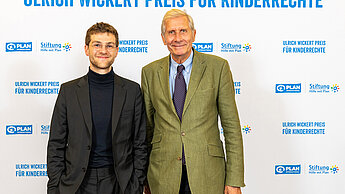 Anton Stanislawski steht neben Ullrich Wickert vor einem Hintergrund, auf dem "Ulrich Wickert Preis für Kinderrechte" steht