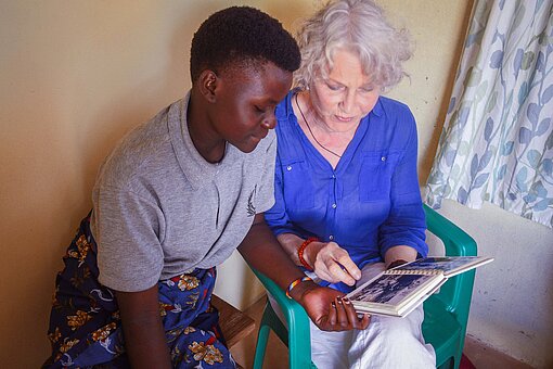 Eine junge Frau aus Tansania sitzt neben einer älteren weißen Frau, sie schauen sich gemeinsam ein Fotobuch an