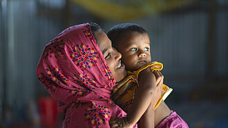 Eine junge Frau mit Kopftuch hält ein Kleinkind auf dem Arm und schaut es lächelnd an