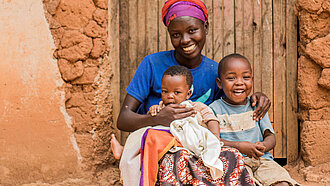 Lächelnde Mutter sitzt vor einer alten Hauswand mit zwei kleinen Kindern auf dem Schoß und neben sich, in traditioneller bunter Kleidung in Ruanda.