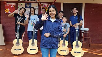 Eine Gruppe Jugendlicher steht draußen, vier von ihnen haben Gitarren vor sich stehen, ein Mädchen steht im Vordergrund und winkt in die Kamera