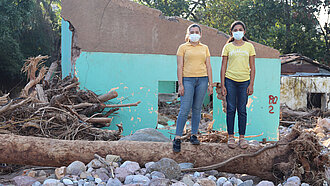 Zwei Mädchen stehen auf einem entwurzelten Baum, der vor einem zertrümmerten Haus liegt.
