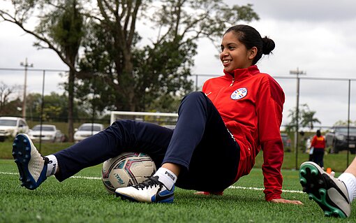 Mädchen in rotem Trainingsanzug sitzt auf dem Fußballrasen und wärmt sich mit einem Fußball auf