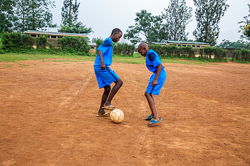 Zwei Jungen, beide in blauem Trikot spielen mit einem Ball auf einem brauen Spielfeld