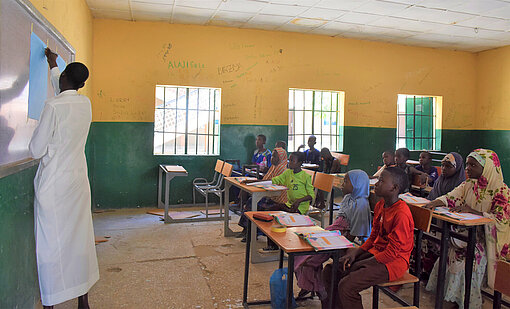 Ein Klassenzimmer mit Schüler:innen in Borno, Nigeria. Ein Lehrer steht an der Tafel und schreibt etwas daran