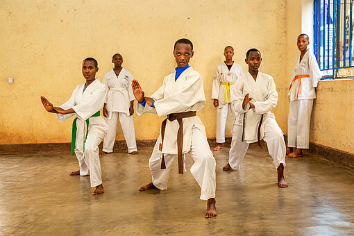 Drei Jugendliche in Karate-Anzügen stehen mit gebeugten Knien und gespannten Armen in einem Raum. Im Hintergrund stehen drei weitere Jugendliche und schauen ihnen zu
