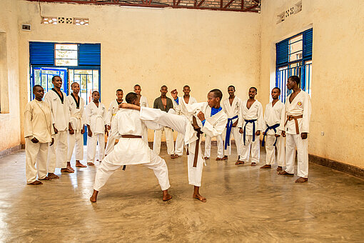 Zwei Jugendliche in Karate-Anzügen kämpfen miteinander. Im Hintergrund stehen weitere Jugendliche im Halbkreis und schauen ihnen zu