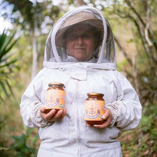 Eine Frau im Imkeranzug steht in einem Wald und hält zwei Honiggläser in den Händen.