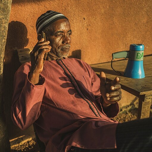 Bamba sitzt mit dem Handy in der Hand vor seiner Hütte