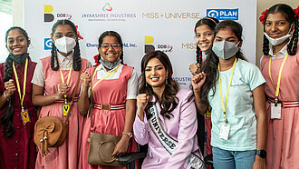 Eine Gruppe Mädchen steht neben einer Frau, die eine Miss Universe-Schärpe trägt und eine Faust für Empowerment ballt