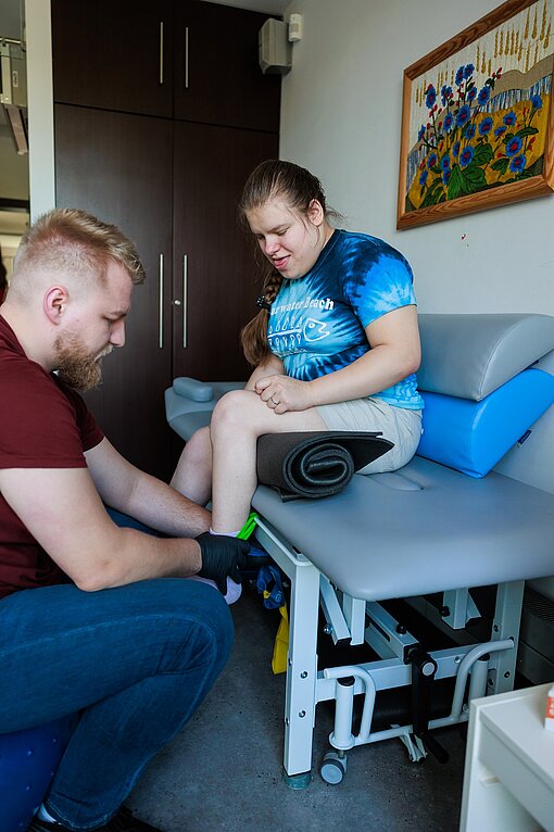 Ein Physiotherapeut behandelt eine junge Patientin, die vor ihm auf einer Liege sitzt, am Fuß.