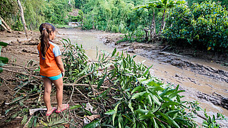 Ein Mädchen steht vor einer schlammigen Straße, die durch Überschwemmungen unbefahrbar geworden ist