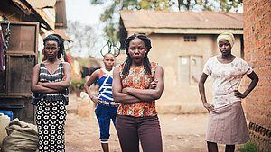Vier junge Frauen stehen mit selbstbewusster Haltung vor einer Häusergruppe und schauen in die Kamera.