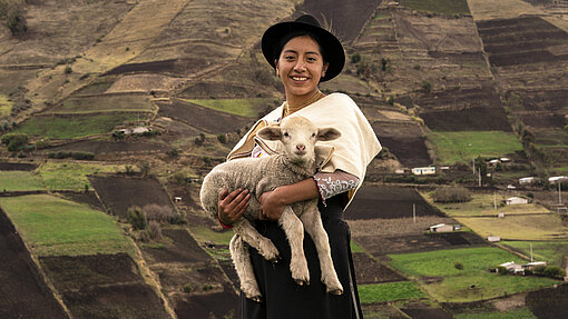 Schafe für berufliche Zukunft junger Menschen in Bolivien. Ein Mädchen steht mit einem Schaf auf dem Arm vor Feldern in Ecuador.