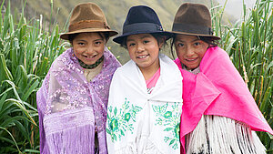 Spenden für Mädchen Drei Mädchen sitzen auf einer Bank. Sie tragen einen Rock, darüber ein Tuch und einen Hut. Sie grinsen in die Kamera. Hinter ihnen ist eine Berglandschaft zu sehen.